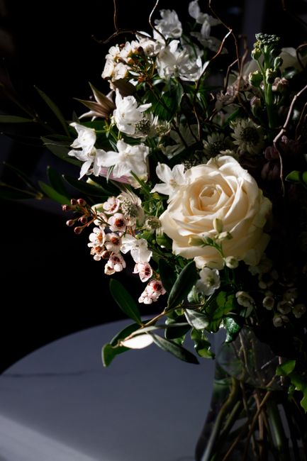 A floral arrangement featuring a large, cream-colored rose at the center, surrounded by clusters of white flowers including smaller roses and delicate blossoms. The bouquet is complemented by green fo
