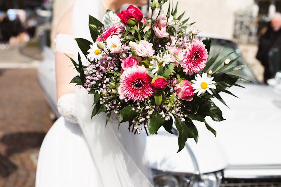 A floral bouquet held by a person dressed in white, featuring vibrant pink gerbera daisies, pale pink roses, white daisies with yellow centers, and delicate pink baby's breath. The arrangement is lush