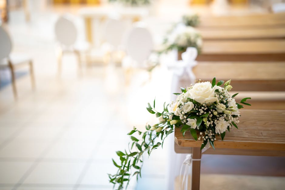 A close-up image of a floral arrangement featuring a bouquet of white roses, lisianthus, and baby's breath, accented with lush green foliage, resting on the edge of a wooden pew at a wedding venue. Th