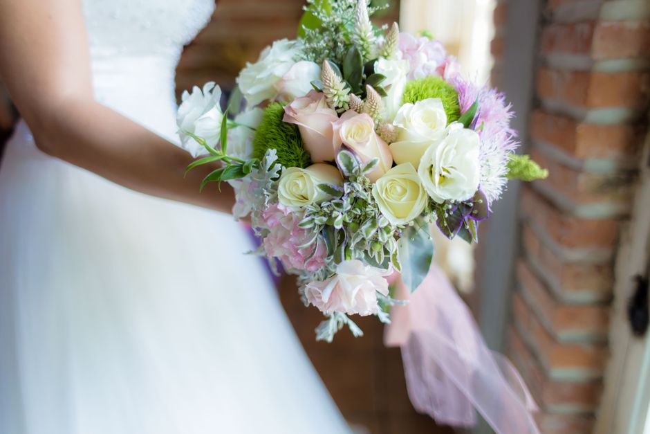 A close-up view of a floral bouquet held by a bride, featuring elegant white roses, pale pink hydrangeas, and soft green foliage arranged in a rounded, lush style. The bouquet is wrapped with a delica