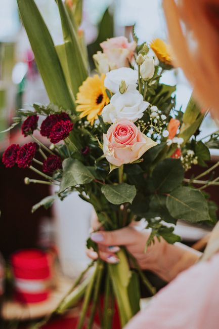 A hand holding a mixed bouquet of fresh flowers, including pale pink roses, white lisianthus, deep red carnations, yellow gerbera daisies, and green foliage, wrapped with clear cellophane and tied wit