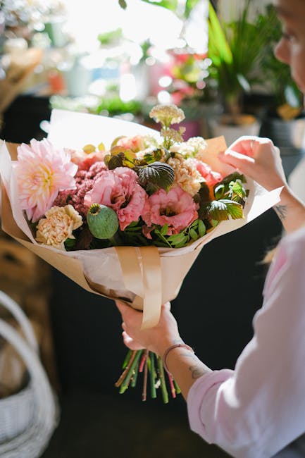 A person holding a large bouquet of fresh flowers arranged with pink and cream peonies, carnations, and delicate white blooms, complemented by lush green leaves. The bouquet is wrapped in white tissue