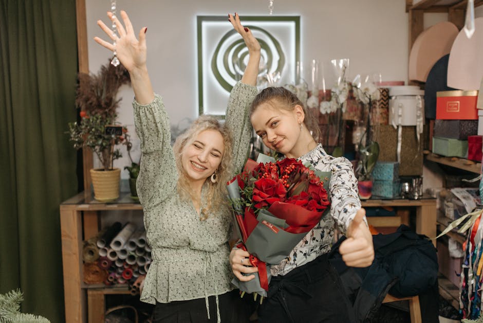 Two young women inside a florist shop are celebrating, one holding a large bouquet of vibrant red roses wrapped in grey and red paper, while the other is smiling broadly with her hands raised in joy. 