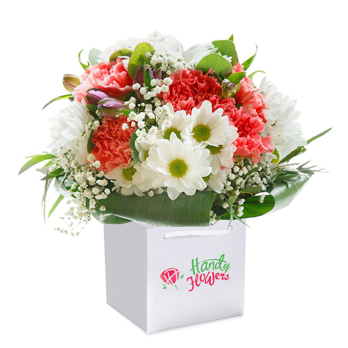 Within a pristine white gift bag, a lush, rounded bouquet stands tall-its composition reflecting the refined craftsmanship of Byfleet florists. Dominating the scene are coral, peach, and vibrant red carnations, each petal intricately ruffled and glowing with lively freshness. Snowy white daisies appear throughout the bouquet, their bright yellow-green hearts recalling the joyful tones of Byfleet's spring wildflowers. Sprigs of delicate white gypsophila create a light, airy filler, lending the arrangement a sense of spaciousness akin to the open greens around Byfleet village. Tucked artfully on the left, subtle purple-tinged flower buds add a dimension of intrigue and promise, their colours hinting at the subtle transitions from winter to spring seen along the edges of Brooklands Park. Encircling broad, shadowy green leaves provide an elegant finish, wrapping the bouquet in a glossy natural frame. White ribbon handles add function and style, making this a perfect choice for easy delivery anywhere across Byfleet-ideal for birthdays, anniversaries, or to show appreciation to someone near High Road or Station Road. Each flower is chosen and arranged with a sense of local insight, showcasing the thoughtful, expert touch for which Byfleet's florists are known.