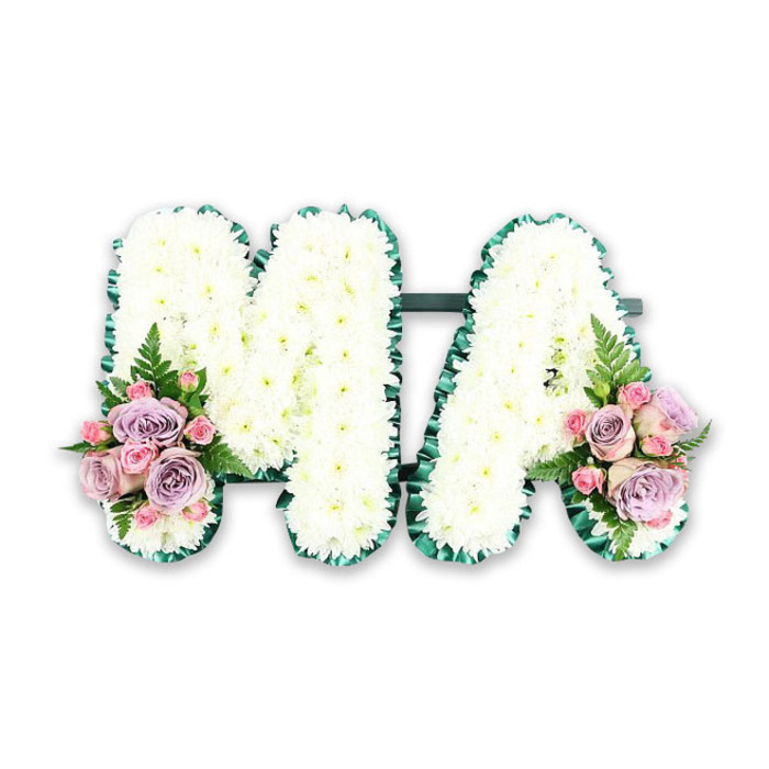 This image presents a personalised funeral letter tribute in the form of the capital letters MA, designed with a balance of structure and tenderness that speaks strongly of family love. Both letters are composed of pure white chrysanthemums, the blooms arranged so densely that their textured centres and fine petals blend into a single, velvety surface. The chrysanthemums are uniform in size and colour, giving the tribute a clean, pristine look, like crisp linen laid out carefully on a table. Around the edges of the 'M' and 'A' runs a framing border of deep green, likely created from folded ribbon or foliage gathered into slight ruffles. This border provides a subtle, sculpted outline, ensuring the letters remain clearly defined whether placed by the casket, near the front of a church, or within a Byfleet funeral home. At the lower left side of the 'M' and the lower right side of the 'A', two graceful mini-bouquets add gentle colour. These posies feature lilac and pale pink roses alongside smaller pink spray roses, each bloom unfurling with velvety petals in soft, romantic hues. Wisps of fern foliage rise from within the roses, their fine, feathery leaves adding height and a touch of wild garden character, reminiscent of the greenery near Byfleet's village lanes and riverside paths. The arrangement is photographed in even, neutral light, with no distractions around it, allowing the viewer to focus on the craftsmanship: the shaping of the letters, the neatness of the edging, the harmony of the colour palette. It is the kind of bespoke floral tribute chosen to honour a mother, mama, or loved one with initials M.A., conveying respect, peace, and enduring affection through flowers rather than words.