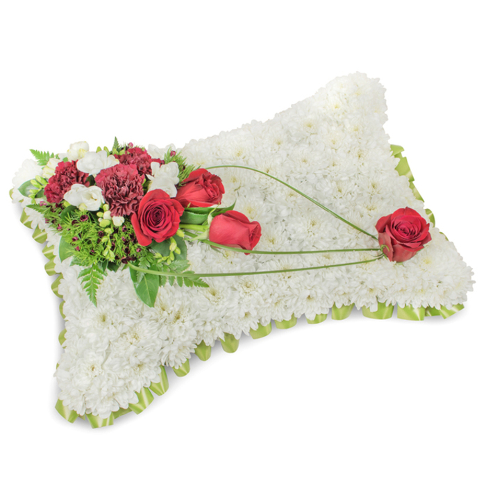 An exquisitely composed funeral pillow, shaped as a gentle rectangle and entirely covered with hundreds of small white chrysanthemums, reads at once as soft, pure, and consoling. The dense white surface has a tactile, almost blanket-like quality that invites the eye to rest, while a faint edge of light green ribbon or foliage peeps between the blooms to define the form with a fresh, understated accent. On the upper left a lush cluster of crimson and burgundy flowers-plush red roses in multiple stages of bloom and ruffled deep red carnations-sits with graceful confidence; tiny white freesias or spray chrysanthemums thread through this cluster, offering delicate points of light. A bed of mixed greenery, from feathery fern fronds to broader glossy leaves, gives the spray depth and a natural, living frame. From that cluster, three long, ribboning strands of bright green bear-grass or similar linear foliage arc gracefully across the chrysanthemum field toward the lower right, their sweeping curve creating motion and a gentle narrative line. At that terminus a single, perfectly formed red rose rests, held like a small, solemn offering-an emblem of remembrance and enduring affection. Soft, neutral lighting emphasises the contrast between the cool white base and the warm crimson accents without harsh shadows. This arrangement would read as a dignified statement at services in the Byfleet area, whether placed at St Mary's or near the quiet lanes off the village green; its textures, colours, and measured composition convey composure, sympathy, and deep respect.